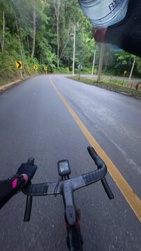 Vertical POV footage of a cyclist holding a water bottle while riding on a wet mountain road after rain, featuring winding curves, tropical forest and endurance cycling.