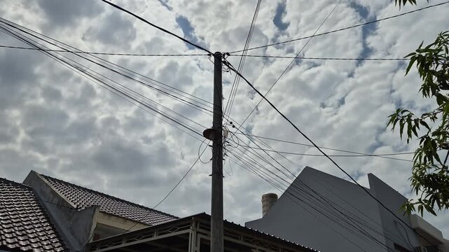 ​Concrete utility pole with tangled power lines and messy electrical cables in a residential neighborhood under a cloudy daytime sky.