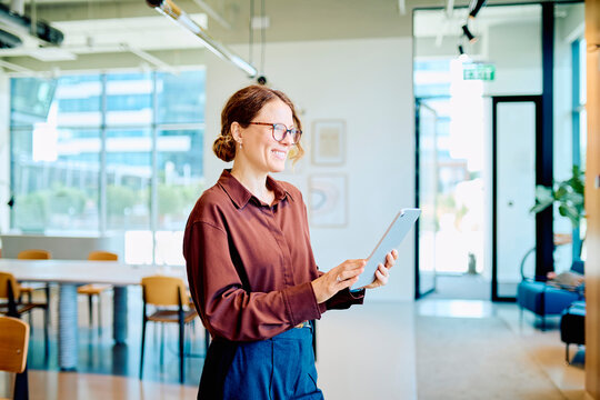 Smiling mid adult businesswoman holding a digital tablet. She is standing in a modern office, engaged with technology and work