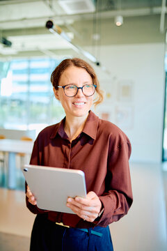 Confident professional woman standing in a modern office, smiling and using a digital tablet for work, embracing technology and business