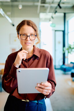 Young professional woman with glasses and earbuds standing in a modern office, working with a digital tablet