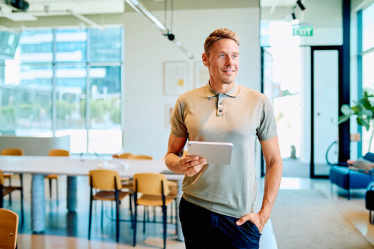 Confident businessman standing in a bright coworking space, holding a digital tablet and looking away, representing innovation and technology
