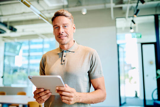 Confident business man smiling while holding a digital tablet in a bright, contemporary workspace, symbolizing technology and success