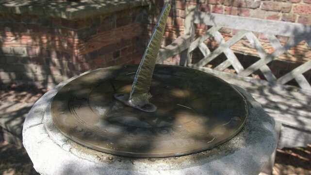 Close up of an ornate historic brass sundial in Well Hall Park, Eltham, with sunlight and tree shadows moving across the engraved dial in a peaceful garden setting.