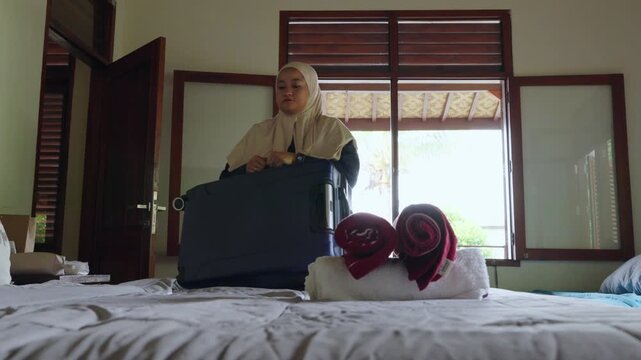 muslim woman wearing hijab packs suitcase beside neatly made bed in the hotel