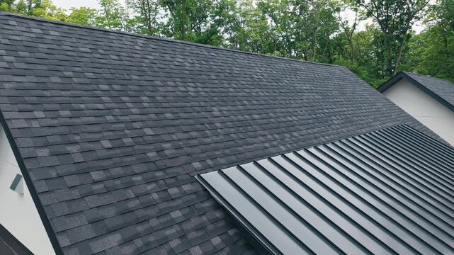 Drone close up view of a modern residential roof of house with a newly installed black asphalt shingle roof surrounded by mature forest. 
