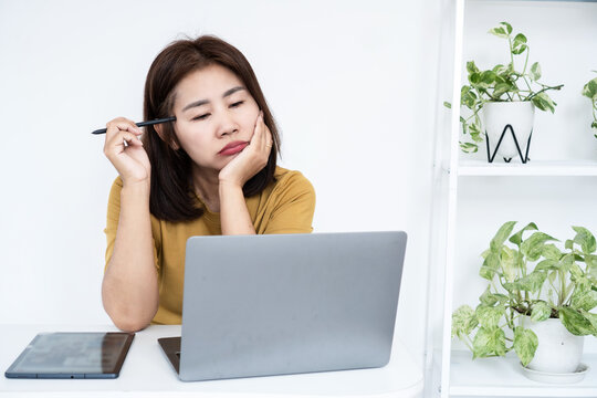 Thoughtful Asian business woman holding stylus pen while working on laptop in home office. Bored or tired freelancer pondering over problem, decision making, or creative block at work desk