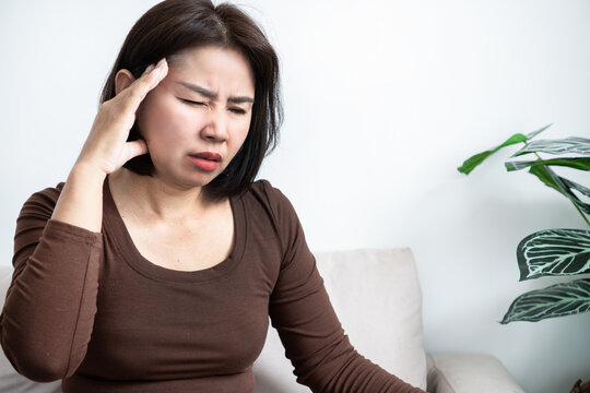Stressed Asian woman suffering from painful headache and dizziness while sitting on couch at home. Exhausted female touching head, feeling migraine symptom, fatigue, or illness