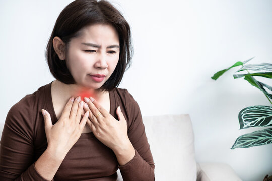Asian woman touching neck suffering from severe sore throat and painful inflammation. Sick female experiencing throat pain, thyroid problem, illness symptom, or difficulty swallowing