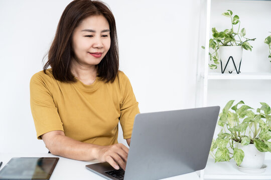 Happy Asian woman smiling while working on laptop computer at home workspace. Cheerful adult female freelancer typing on keyboard with tablet