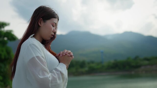 Faith and prayer concept. Young Asian woman with long hair wearing white dress clasping hands in prayer by the lakeside with scenic green mountains and water background. Spiritual peace, hope.