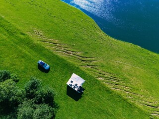 Obraz premium Aerial view of green grass field with a car and tent by a calm lake under clear blue sky