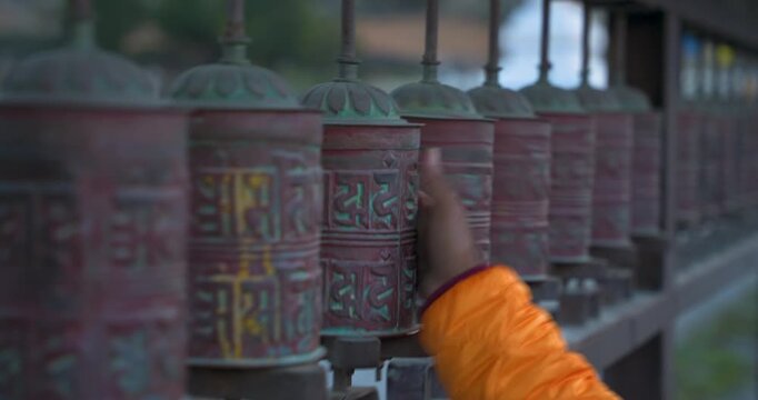 Annapurna Circuit, Manang, Nepal, close-up camera shot of a traveler in an orange jacket spinning a row of traditional bronze Buddhist prayer wheels.