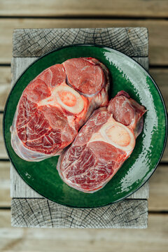 Top view of two raw cross-cut beef shanks with exposed marrow bones arranged on a glossy green plate over a rustic wooden surface