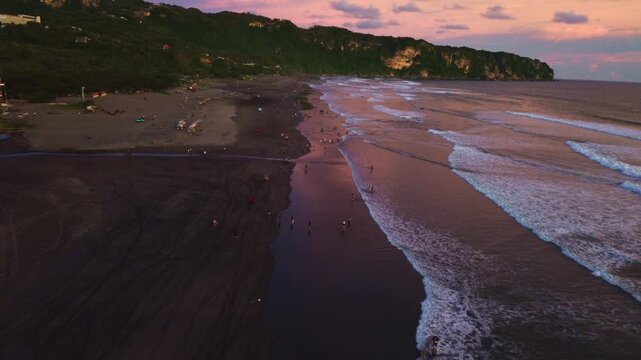 Sweeping aerial drone shot of people enjoying a vibrant sunset on Parangtritis Beach, Yogyakarta, with waves and dramatic cliffs.