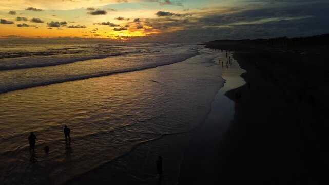 Cinematic aerial drone shot of silhouetted people enjoying a vibrant golden hour sunset on Parangtritis Beach, Yogyakarta, with gentle ocean waves.