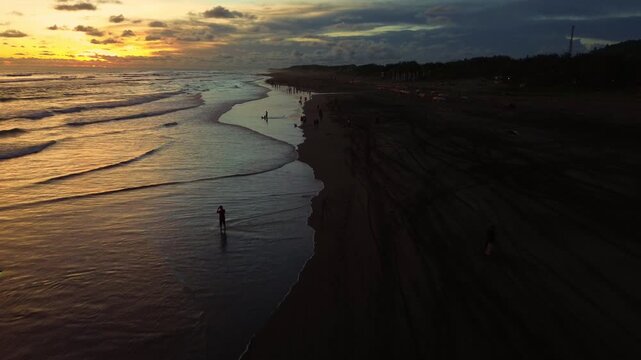 Cinematic aerial drone shot of people enjoying the vibrant golden hour sunset at Parangtritis Beach, Yogyakarta, with waves gently crashing.