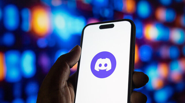 Person holding smartphone with Discord application logo on screen against blurry bokeh background.