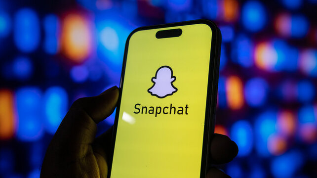 Snapchat application logo displayed on a smartphone screen held by a hand with blurred colorful background lights.