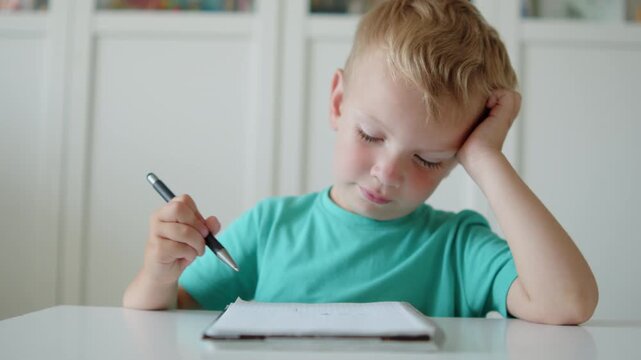 Bored boy writing in a notebook with a pen. Tired little child studying in class. Depressed kid writing in notebook at desk in school or kindergarten. Sad baby student scribbling in a book.