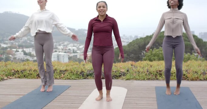 Diverse female friends on yoga mats following center lead, doing jumping jacks on deck, warming up