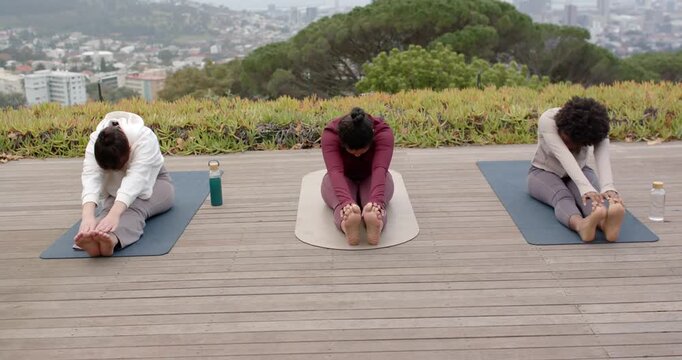 On instructor cue, three women in leggings practicing forward fold on mats, holding feet, rising up