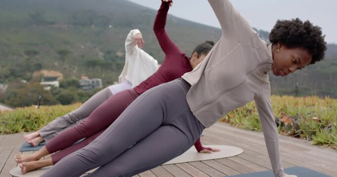 Diverse female friends performing side plank on yoga mats on deck, front leading torso rotations