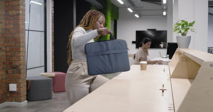 African American woman entering with cup and navy bag, unzipping, opening laptop to work at counter