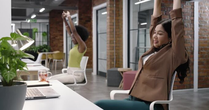 Diverse women staff on break, stretching and rolling necks, easing tension at office with laptop