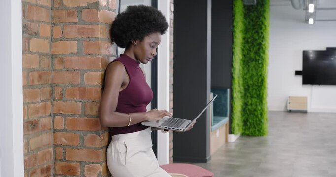 African woman in maroon top leaning in office listening on earbud and typing laptop answering call