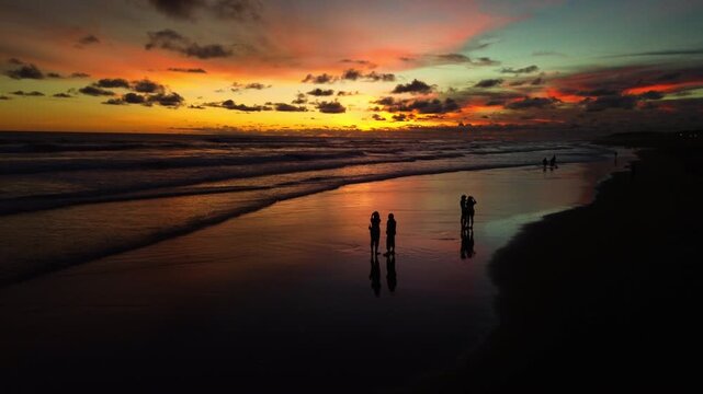 Cinematic wide shot of people silhouettes walking and enjoying a vibrant sunset on Parangtritis Beach, Yogyakarta, with ocean waves reflecting the colorful sky.