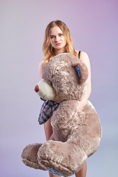 A slender young woman with an elegant figure is hugging a huge brown teddy bear. She is wearing black lingerie. The bear has a white nose and a blue earband.