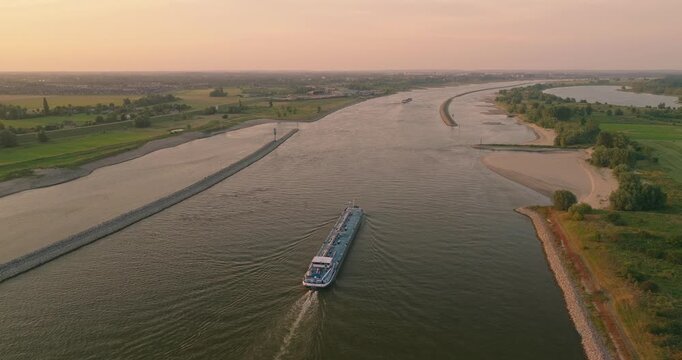 Beautiful drone shot of a cargo ship sailing through a Dutch canal at sunset, surrounded by the iconic polder landscape and vibrant colorful skies over the peaceful countryside.