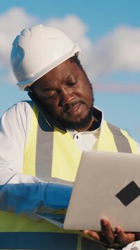 Multitasking engineer or foreman in construction site, vertical portrait of black man with laptop and mobile phone outdoors. Negotiating with contractors by phone, architecture project implementation