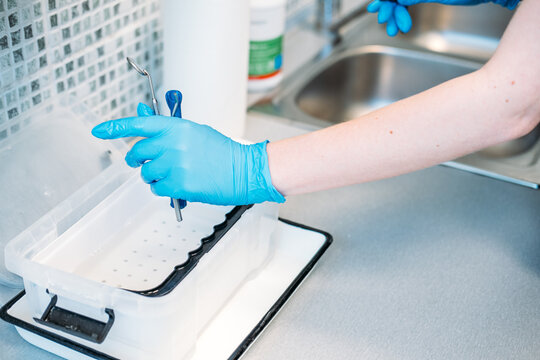 Gloved hand places dental tools into plastic disinfection container near sink. Dental instrument soaking, disinfectant solution tray, presoak cleaning dental tools.