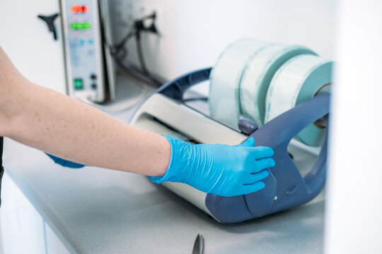 Gloved hand operates sealing machine with rolls of sterilization pouches on counter. Sterilization pouch roll, sealed sterile packaging, medical instrument packaging.