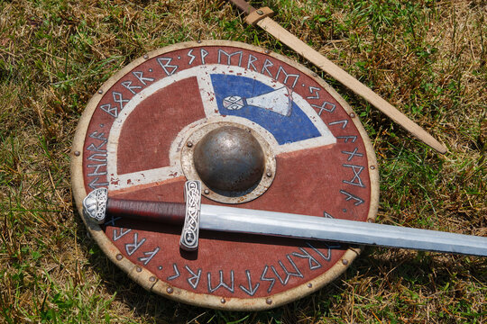 Authentic looking Viking age weaponry including a round shield and a long sword are displayed on a green grassy field for historical reenactment.