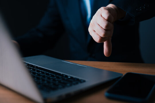Businessman showing thumbs down gesture in dark office. Concept of rejection, failure, bad review, disapproval, or poor performance. Male hand expressing negativity near laptop.
