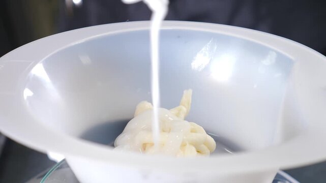 Side close-up of white creamy hair dye being squeezed from white bottle into clear plastic bowl. Smooth texture, bright light reflections, and clean product preparation for salon coloring 4k video