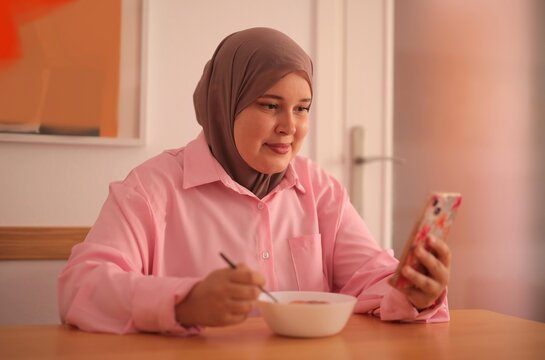 Woman in hijab scrolls on phone while eating breakfast