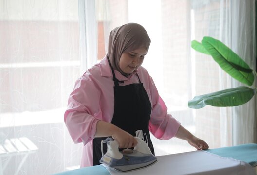 Muslim woman ironing clothes next to a bright window
