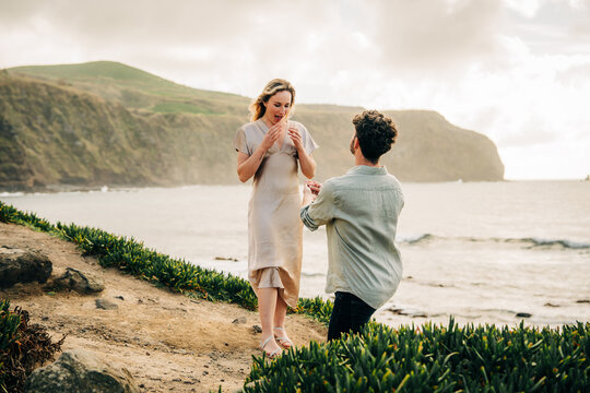 Man on one knee proposes to surprised woman by the sea
