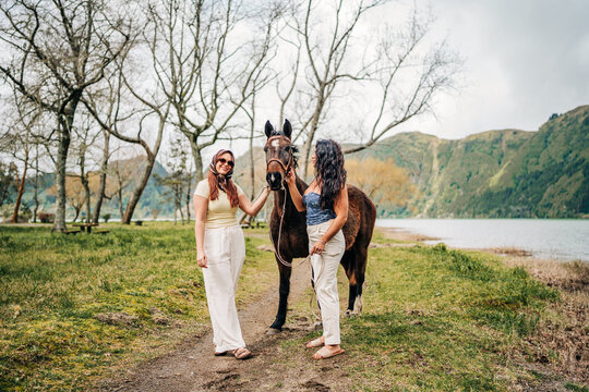 Two stylish women friends petting a horse by a lake