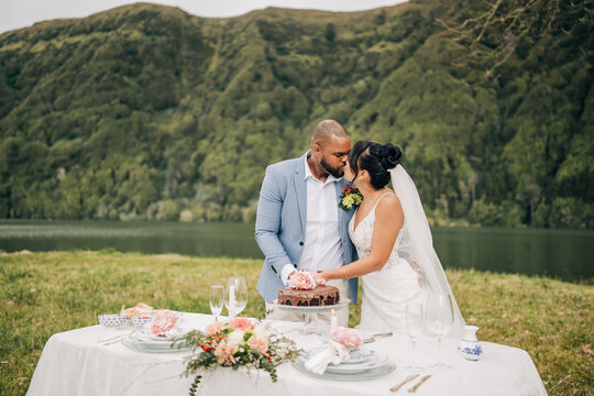 Bride and groom kiss cutting cake by mountain lake