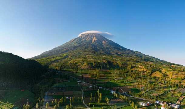 Aerial view panoramic of Mount Sindoro volcano with lush green agricultural terraced fields, scenic village settlement, and a clear blue sky in Wonosobo, Central Java, Indonesia.