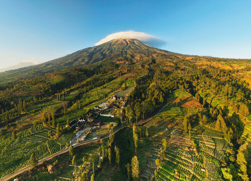Aerial view panoramic of Mount Sindoro volcano with lush green agricultural terraced fields, scenic village settlement, and a clear blue sky in Wonosobo, Central Java, Indonesia.