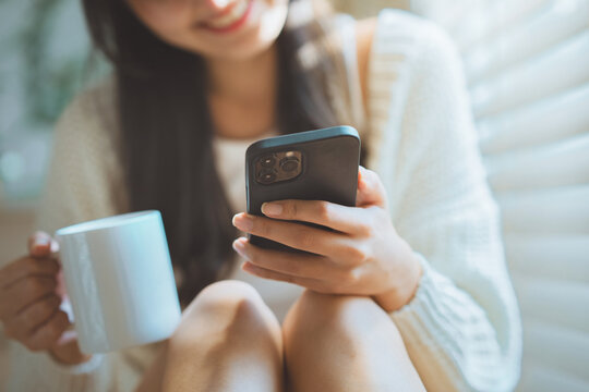 Close up of Asian woman holding coffee mug and using smartphone by window with sunlight, happy female smiling while texting on mobile phone online app at home bedroom for cozy leisure, digital lifesty