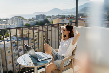 Obraz premium Millennial woman working on laptop on sunny apartment balcony with summer city view.