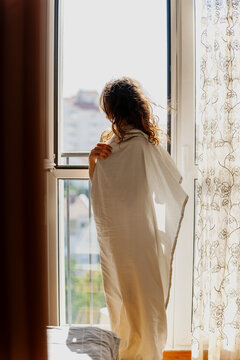 Beautiful young woman standing by window wrapped in white sheet, greeting a new day and looking at the city.