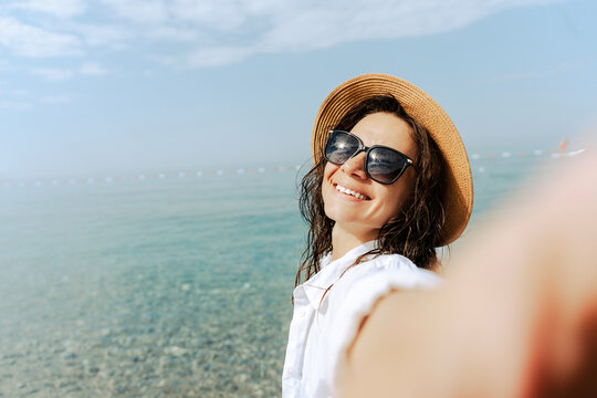 Happy young woman in summer hat and white shirt taking a selfie on sea beach.